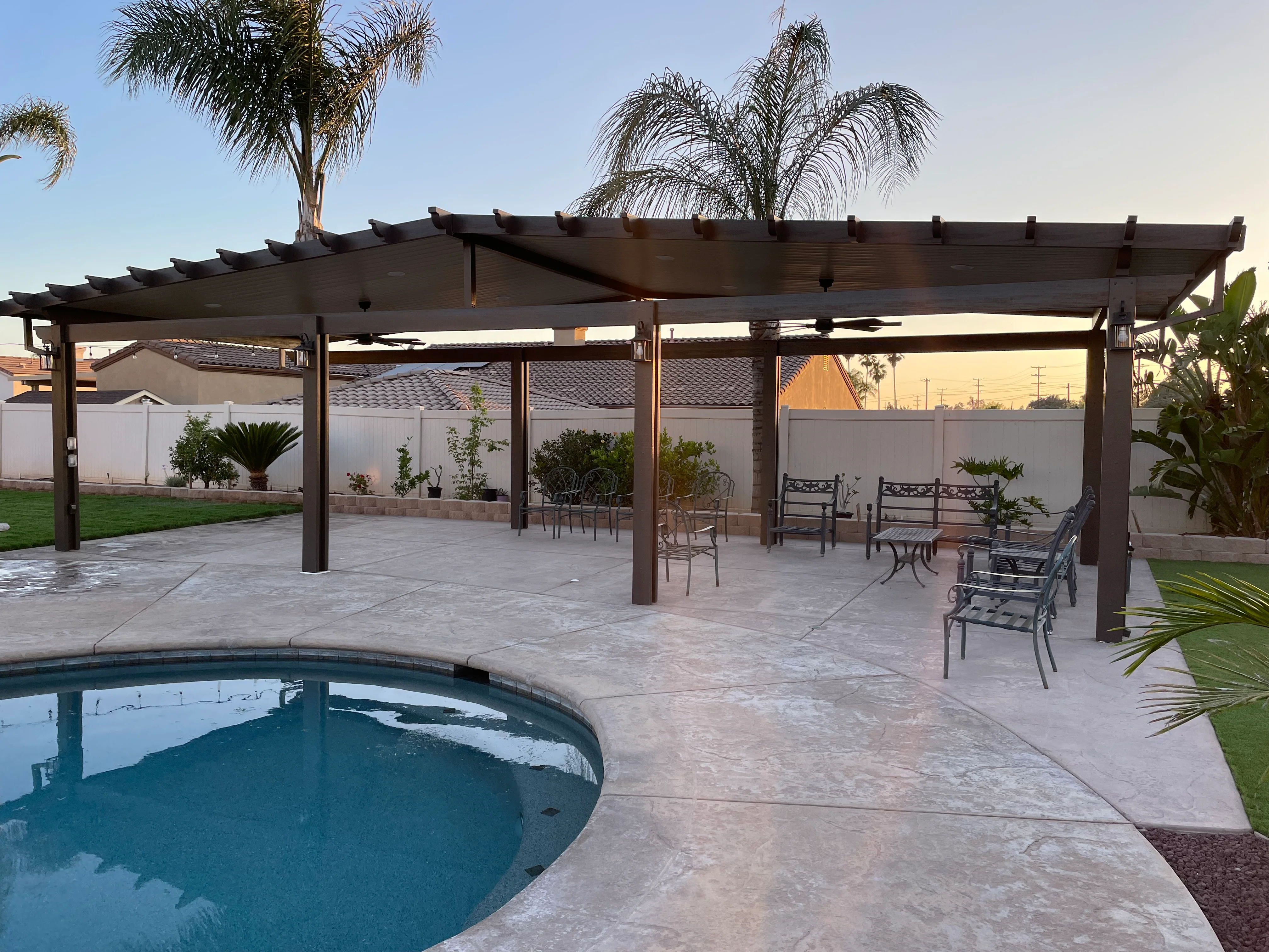 Dark wood pergola over concrete patio with pool, metal seating, and palm trees