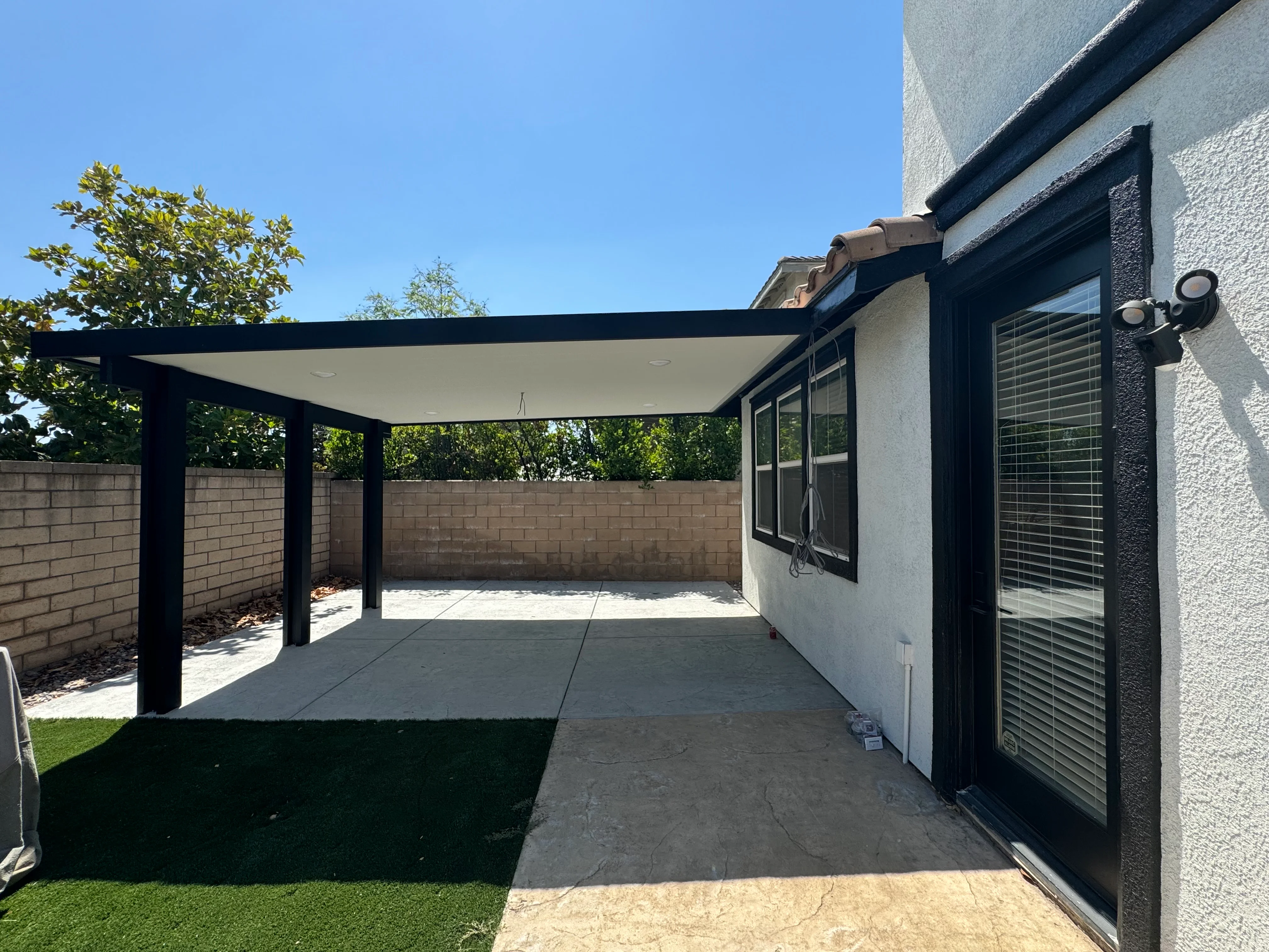 Black pergola with white shade canopy over concrete patio beside modern home
