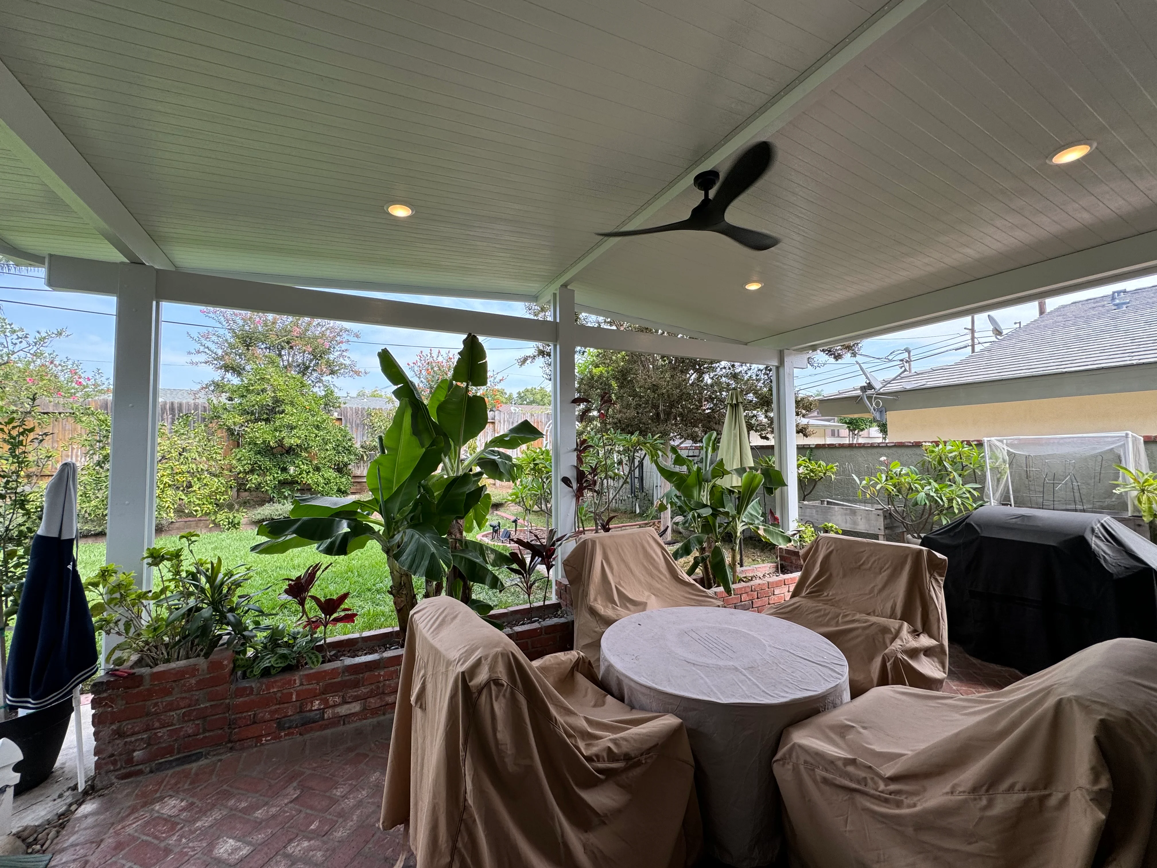 Covered patio with white soffit ceiling, ceiling fan, recessed lights, and brick planter boxes with tropical plants