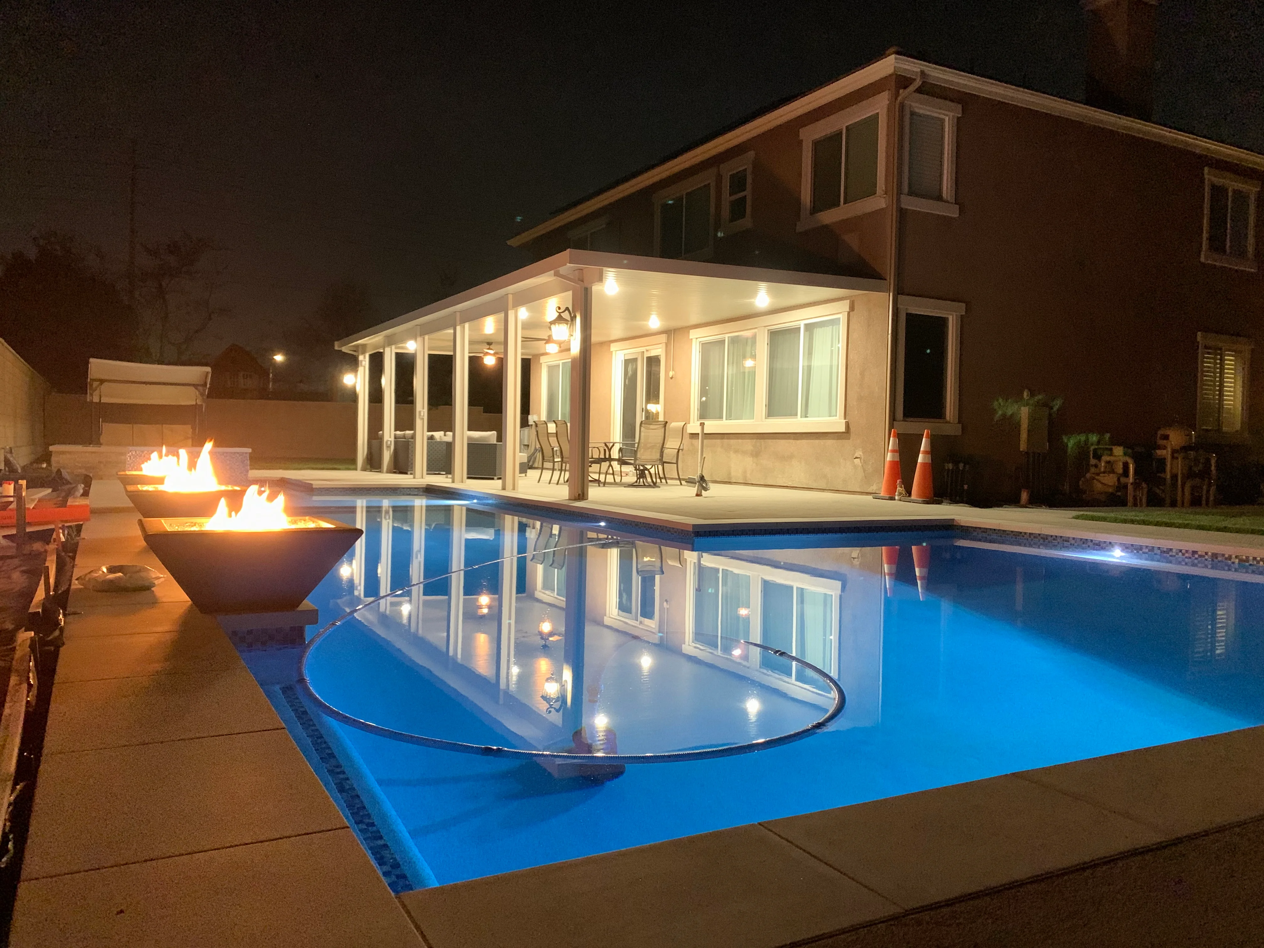 Nighttime view of illuminated pool with covered patio, fire features, and modern home